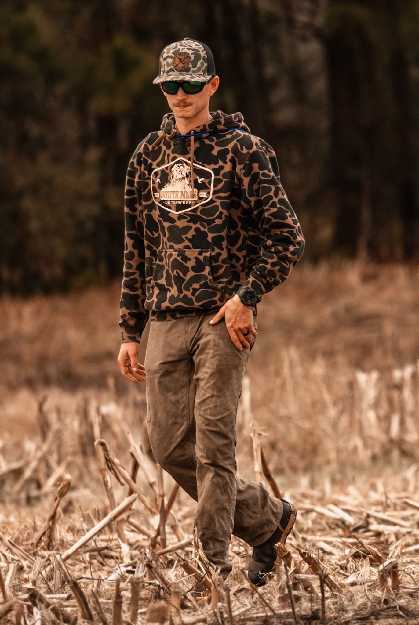 Man wearing South Neck duck camo hoodie walking through harvested field – outdoor lifestyle and hunting apparel for men