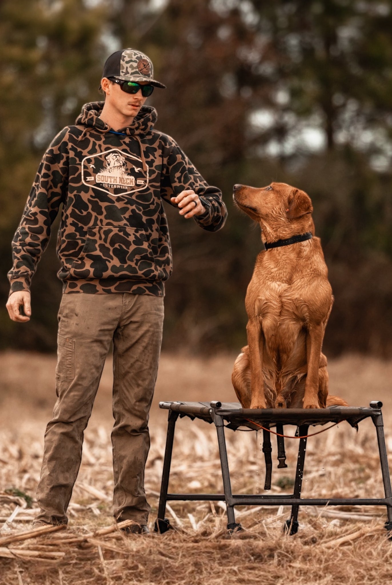 Man wearing South Neck duck camo hunting hoodie training a Labrador retriever in a field – outdoor hunting apparel for dog trainers and sportsmen